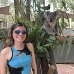 A person wearing sunglasses and a tie-dye tank top stands smiling next to a koala perched on a tree branch. The setting is outdoors with greenery and eucalyptus leaves in the background.