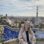 A woman sits on a colorful mosaic bench, with a cityscape and distinctive architectural structures in the background. The sky is cloudy, and she is wearing a light coat. The scene suggests a park or scenic viewpoint.