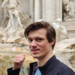 A person is standing in front of the Trevi Fountain in Rome, looking at the camera with a slight smile and a fist raised. Sculptures of mythological figures and flowing water are visible in the background.