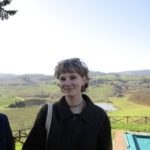 A person with short hair, wearing a dark jacket and sunglasses on their head, stands outdoors holding a drink. They smile, surrounded by a scenic landscape of rolling hills, vineyards, and a clear sky. A pool and a wooden fence are visible nearby.
