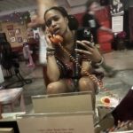 A person sits on the floor, holding a retro-style phone and taking a selfie in a mirror. They are in a room with colorful decor and blurred people in the background. The atmosphere is casual and lively.