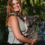 A person with long brown hair, wearing a green sleeveless top and black pants, is smiling while holding a koala. They are outdoors with greenery and water in the background.