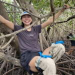 Person sitting and smiling among dense mangrove branches, wearing a black t-shirt and shorts, with protective coverings on their legs secured with blue ties. Another person is partially visible in the background. The setting is lush and green.