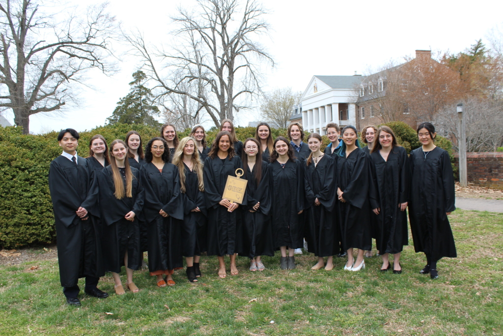 A group of people wearing black graduation gowns stand outdoors on grass, posing for a photo. One person in the center holds a gold book titled "Honor." Leafless trees and a large building with white pillars are in the background.