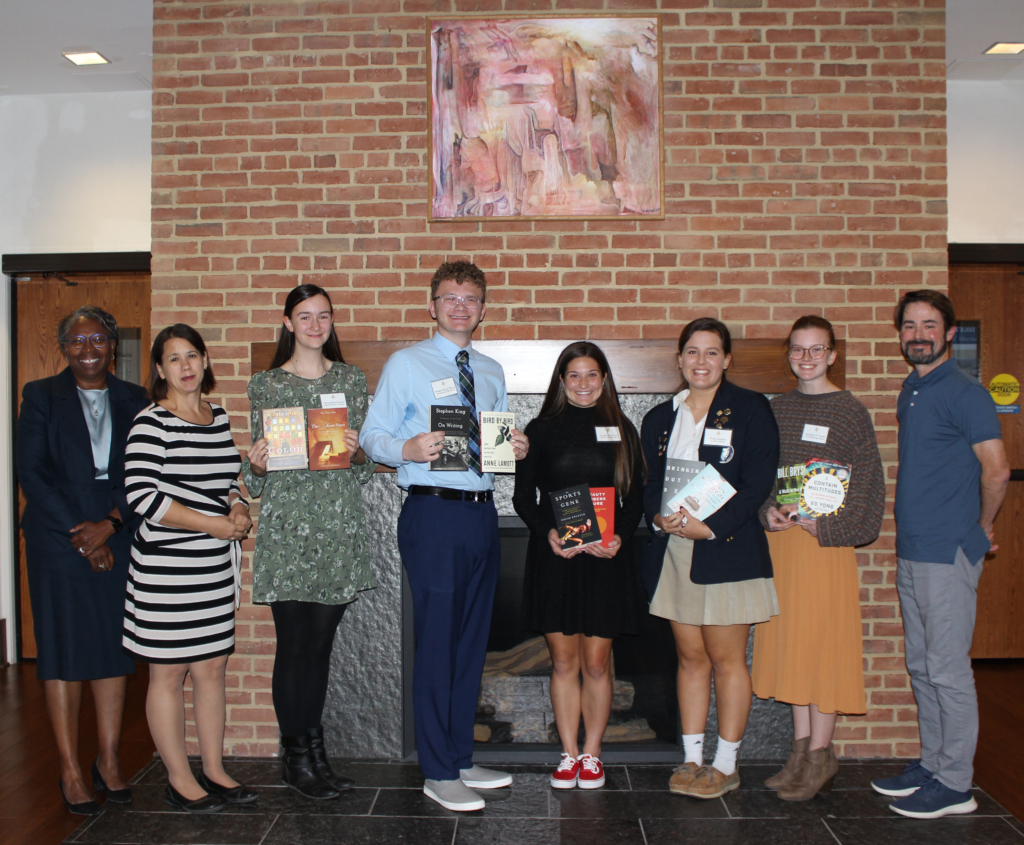 Seven people stand smiling in front of a brick wall, holding books. They are dressed in formal and semi-formal attire. An abstract painting is mounted on the wall behind them.