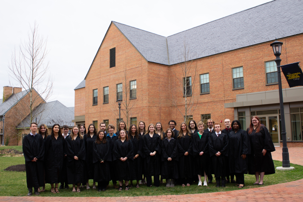 A group of graduates in black gowns stand in front of a brick building with a sloped roof. The scene is outdoors on a cloudy day. There’s a lamppost and a campus sign on the right. Everyone is smiling at the camera.