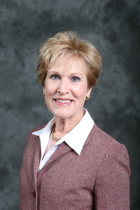 Woman with short blonde hair wearing a light maroon blazer over a white collared shirt, smiling in front of a gray mottled background.