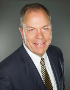 A middle-aged man in a navy suit, white shirt, and striped tie smiles at the camera against a plain gray background.