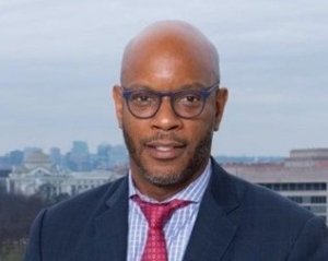 A man wearing glasses, a suit, and a red tie stands outdoors with city buildings visible in the background.