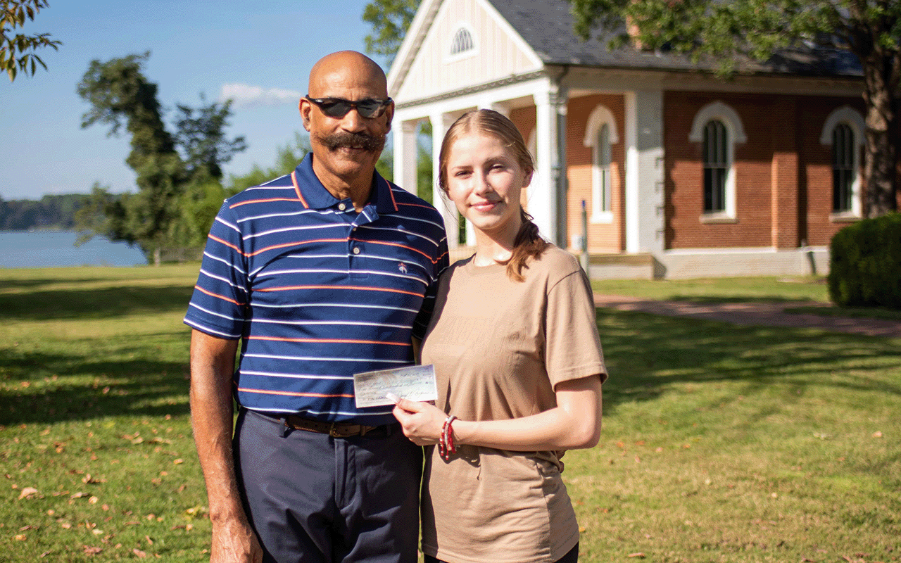 A man and a young woman stand outside holding a check. The man wears sunglasses and a striped polo shirt. The woman wears a brown T-shirt and smiles. A brick building and trees are in the background.