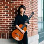 Meng Su standing against brick wall holding her guitar.