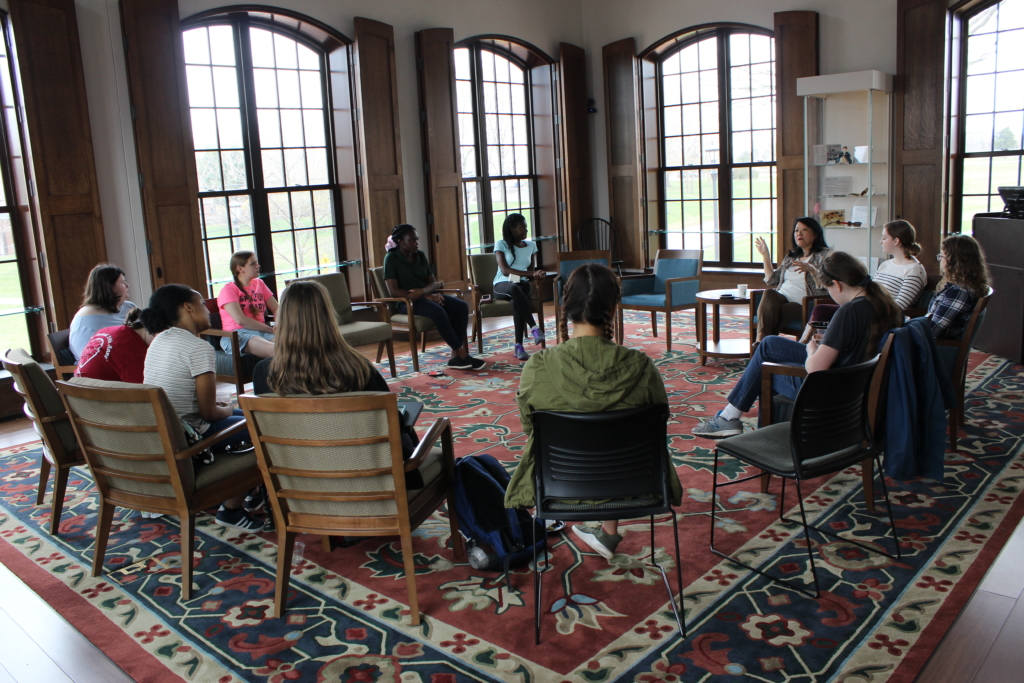 A diverse group of people sit in a circle on chairs in a room with large windows and wooden paneling. They appear to be engaged in a discussion or meeting. The floor features a colorful patterned rug.