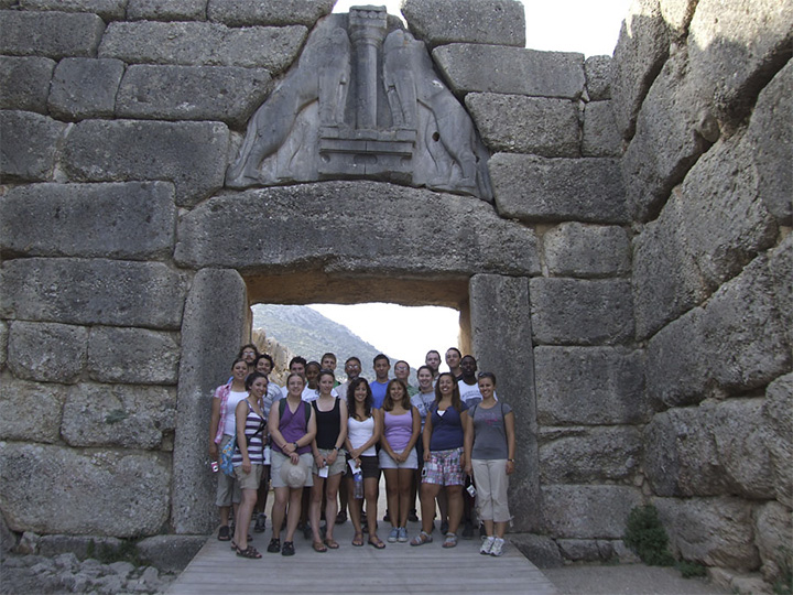 A group of students stand beneath a stone arch with two lionesses carved within.