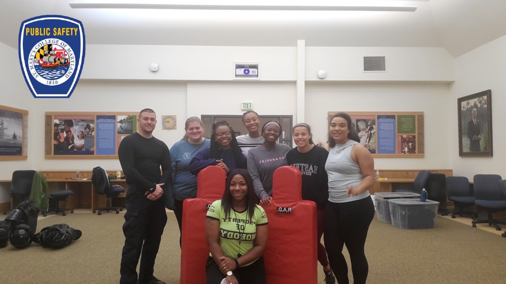 A group of eight people, seven standing and one kneeling, are in a room with educational displays on the walls. They are posing around a pair of red padded training equipment. They appear to be in a classroom or training facility.