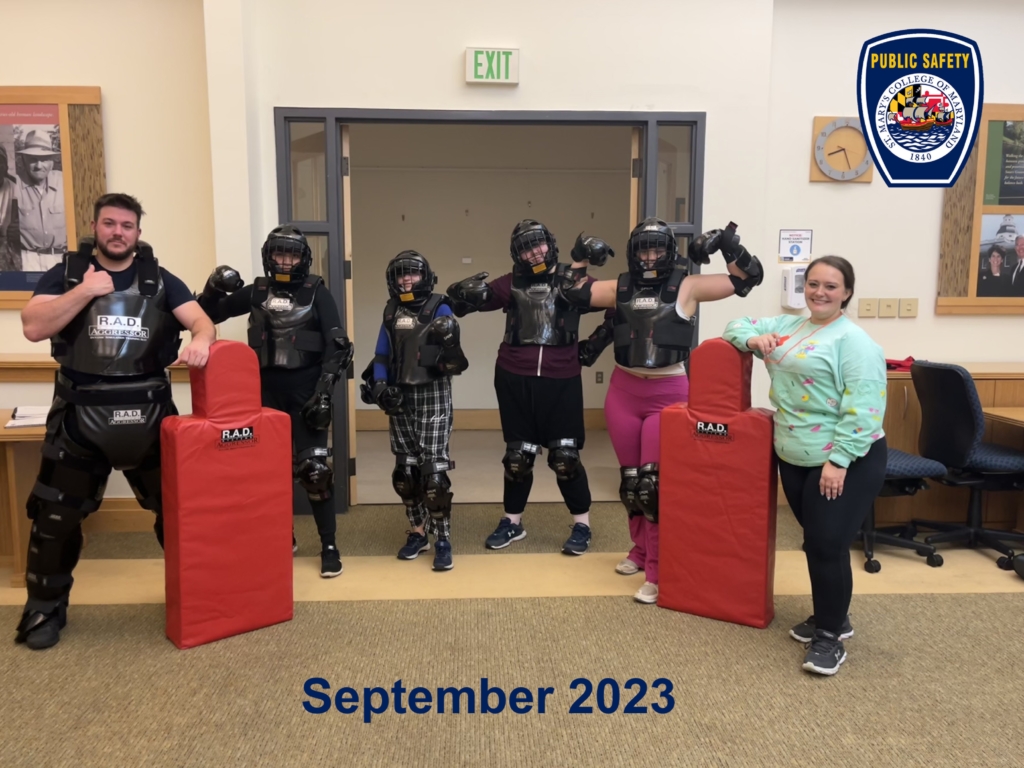 A group of five people wearing protective gear pose with a woman by two red pads. They stand in a room with a sign reading "Public Safety" on the wall. Text at the bottom reads "September 2023.