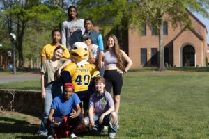 Eight people, including an Seahawk mascot in a 'Mary' jersey, pose on grass with a brick building and trees in the background.