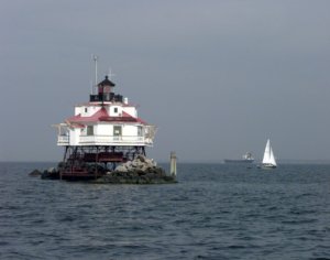 a white lighthouse in the middle of a bay, with a sailboat floating past