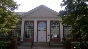 A red brick building with large arched windows and a white triangular pediment. The entrance has double doors and is accessed via a set of stone steps. Surrounded by trees, the building is seen under a clear, bright sky.