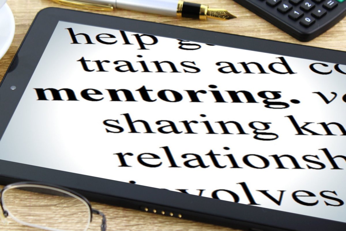 Close-up of a tablet screen displaying large, bold text focused on the word "mentoring." Nearby, a pair of reading glasses and part of a calculator are visible on a wooden table surface.