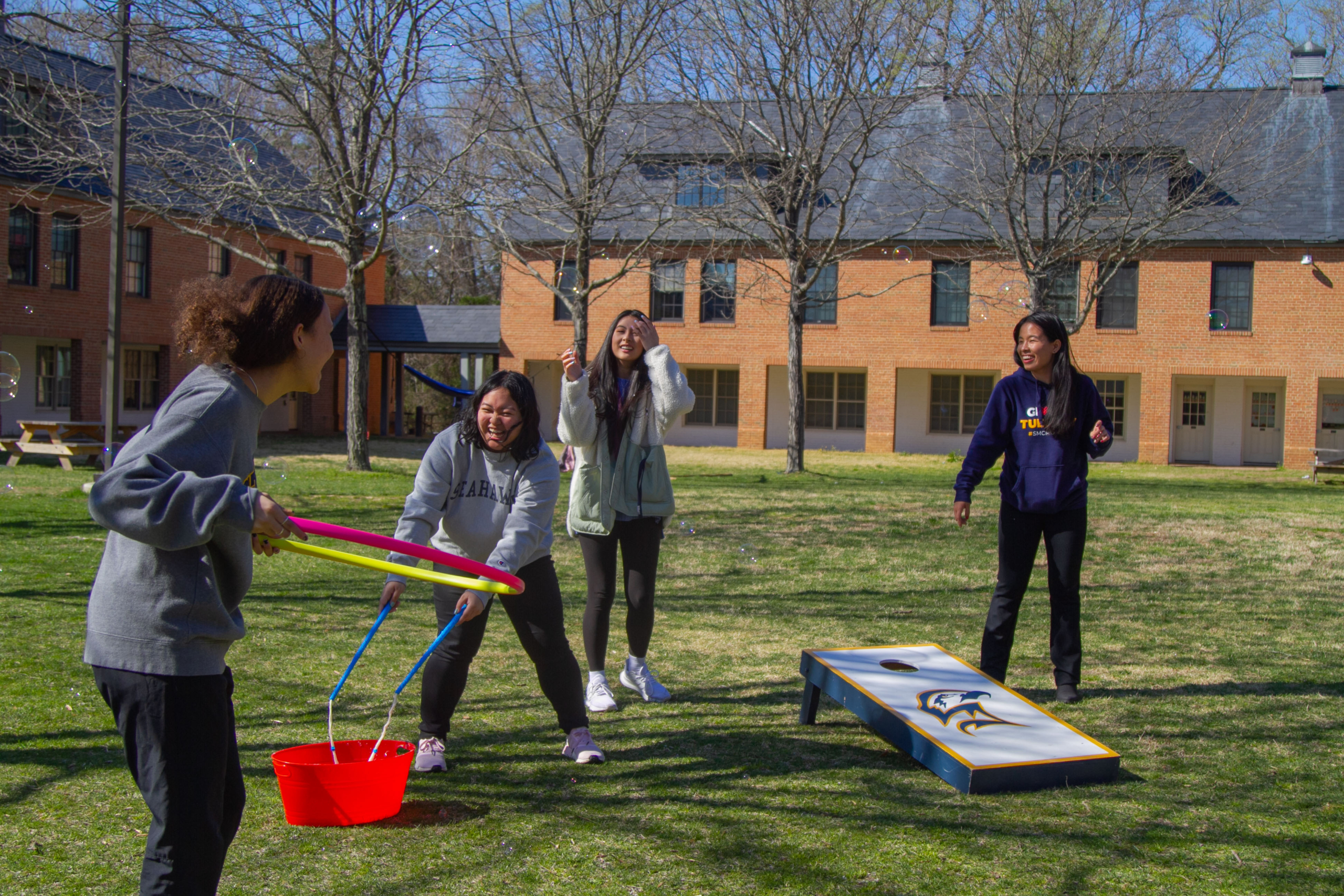 Four female students partake in fun activities like hula hoops, bubbles, and corn hole on the college lawn in front of a residential building