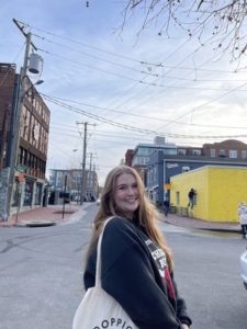 A person with long hair smiles at the camera while standing on a street. They are wearing a dark sweater and carrying a white tote bag. The street is lined with buildings, including a bright yellow one. The sky is overcast.