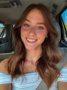 A woman with long brown hair is smiling while sitting in a car. She is wearing a white top and a seatbelt. Sunlight is softly illuminating her face, highlighting her natural makeup and features. The car interior is partially visible.