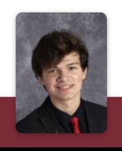 A young person with medium-length brown hair is wearing a black suit, black shirt, and red tie. They are smiling slightly, posed against a gray marbled background.