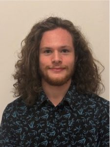 A person with long curly hair and a beard is smiling slightly at the camera. They are wearing a dark shirt with a pattern of light blue leaves. The background is a plain light color.