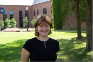 A person with short brown hair wearing a black shirt and necklace stands smiling in a grassy area with trees and a brick building in the background on a sunny day.